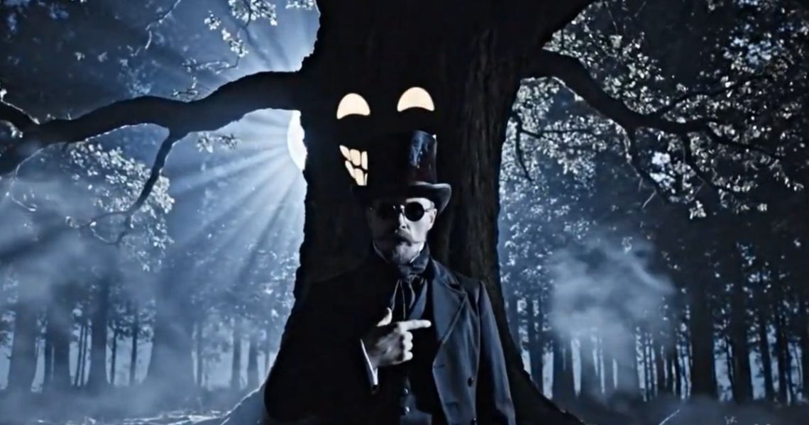 A man with dark glasses and a top hat, standing before a tree which has a face