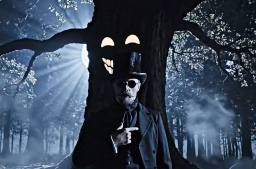A man with dark glasses and a top hat, standing before a tree which has a face
